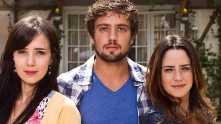 Três pessoas estão alinhadas lado a lado, olhando para a câmera. À esquerda, uma mulher de cabelos escuros e lisos. No centro, um homem de barba curta e cabelos cacheados, usando camisa xadrez aberta sobre uma camiseta azul. À direita, uma mulher de cabelos castanhos claros e ondulados