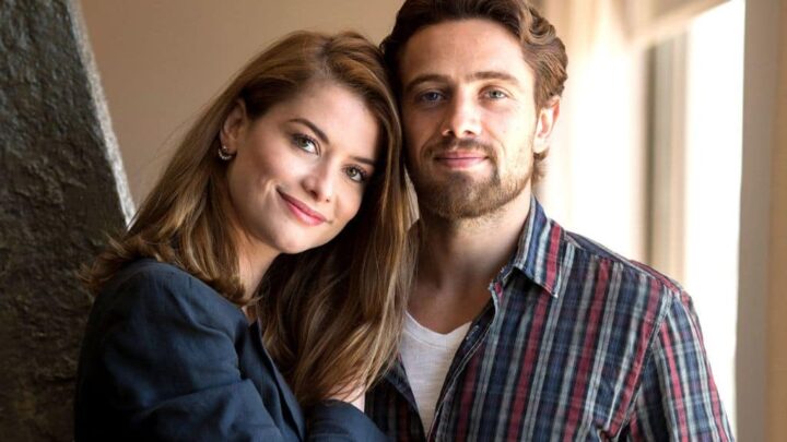 Uma mulher e um homem estão próximos, sorrindo suavemente. A mulher tem cabelos castanhos claros, lisos e veste um blazer azul escuro. O homem tem cabelos castanhos, barba e usa uma camisa xadrez com tons de vermelho e azul