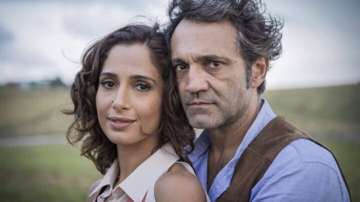 Um homem e uma mulher estão lado a lado em um campo aberto. A mulher tem cabelos cacheados curtos e sorri levemente, enquanto o homem, de cabelos grisalhos e expressão séria, olha diretamente para a câmera