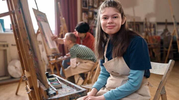 Retrato de uma adolescente com deficiência sorrindo para a câmera sentada perto do cavalete na aula de arte com espaço para cópia.