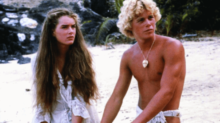 Cena do filme A Lagoa Azul (1980). Um jovem de cabelos cacheados loiros, pele bronzeada e usando apenas uma tanga branca e um colar de concha está ao lado de uma jovem de longos cabelos castanhos, vestindo um vestido branco rústico. Ambos estão em uma praia de areia clara, cercados por rochas e vegetação tropical, olhando para algo fora do quadro com expressões sérias