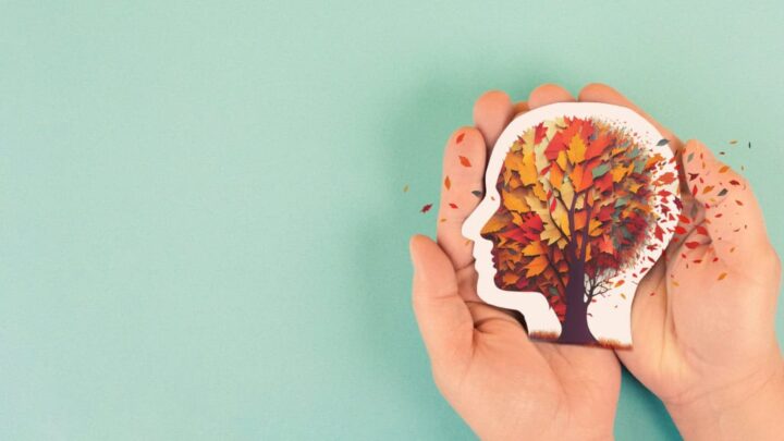 Duas mãos seguram um recorte em forma de cabeça humana, com uma árvore colorida dentro, cujas folhas parecem se desprender. O fundo verde claro destaca a ideia de memória se esvaindo, simbolizando o impacto do Alzheimer.